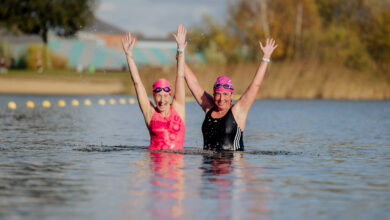 Twee deelnemers nemen een winterdip tijdens een sportevenement bij een recreatieplas, met bomen en een paviljoen op de achtergrond.